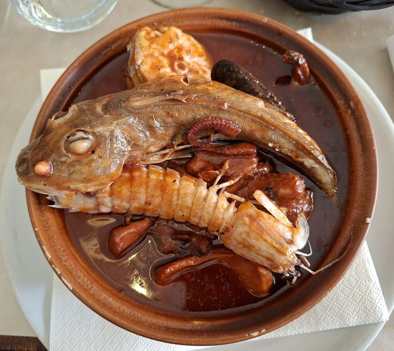 Porzione di cacciucco alla livornese in una ciotola di coccio: pesce intero, canocchia, cozze e polpo nel sugo di pomodoro