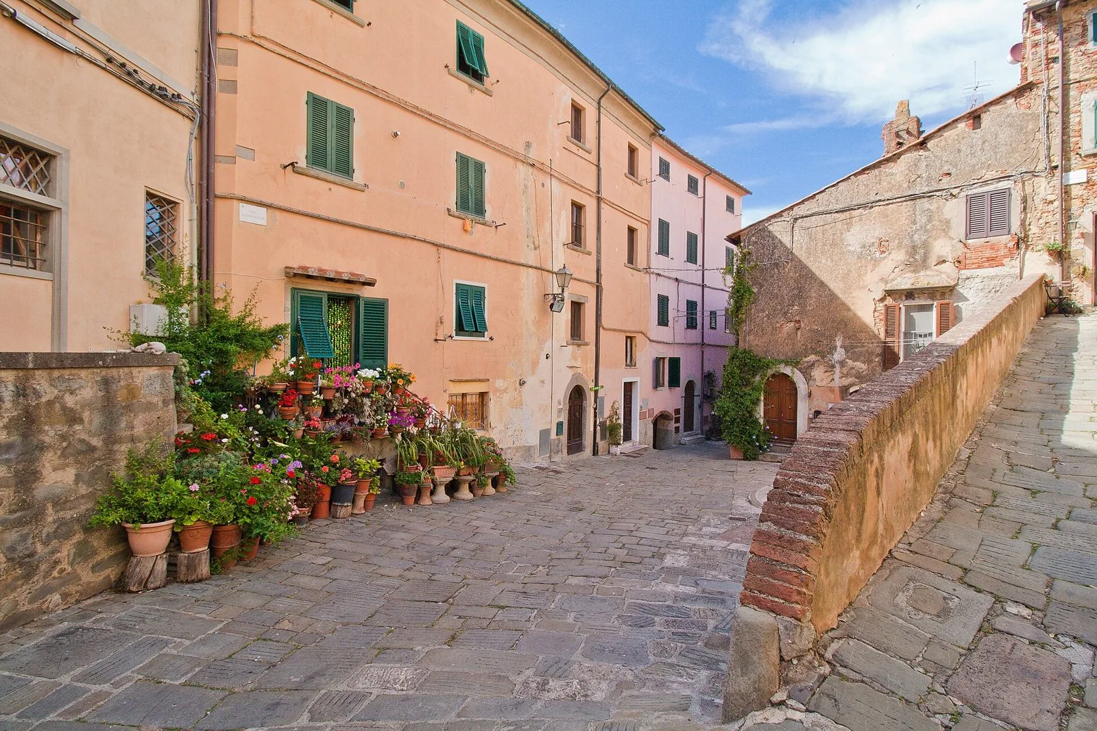 Scorcio del centro storico di Castagneto Carducci: piazzetta lastricata in pietra, case color pesca con persiane verdi e vasi di fiori