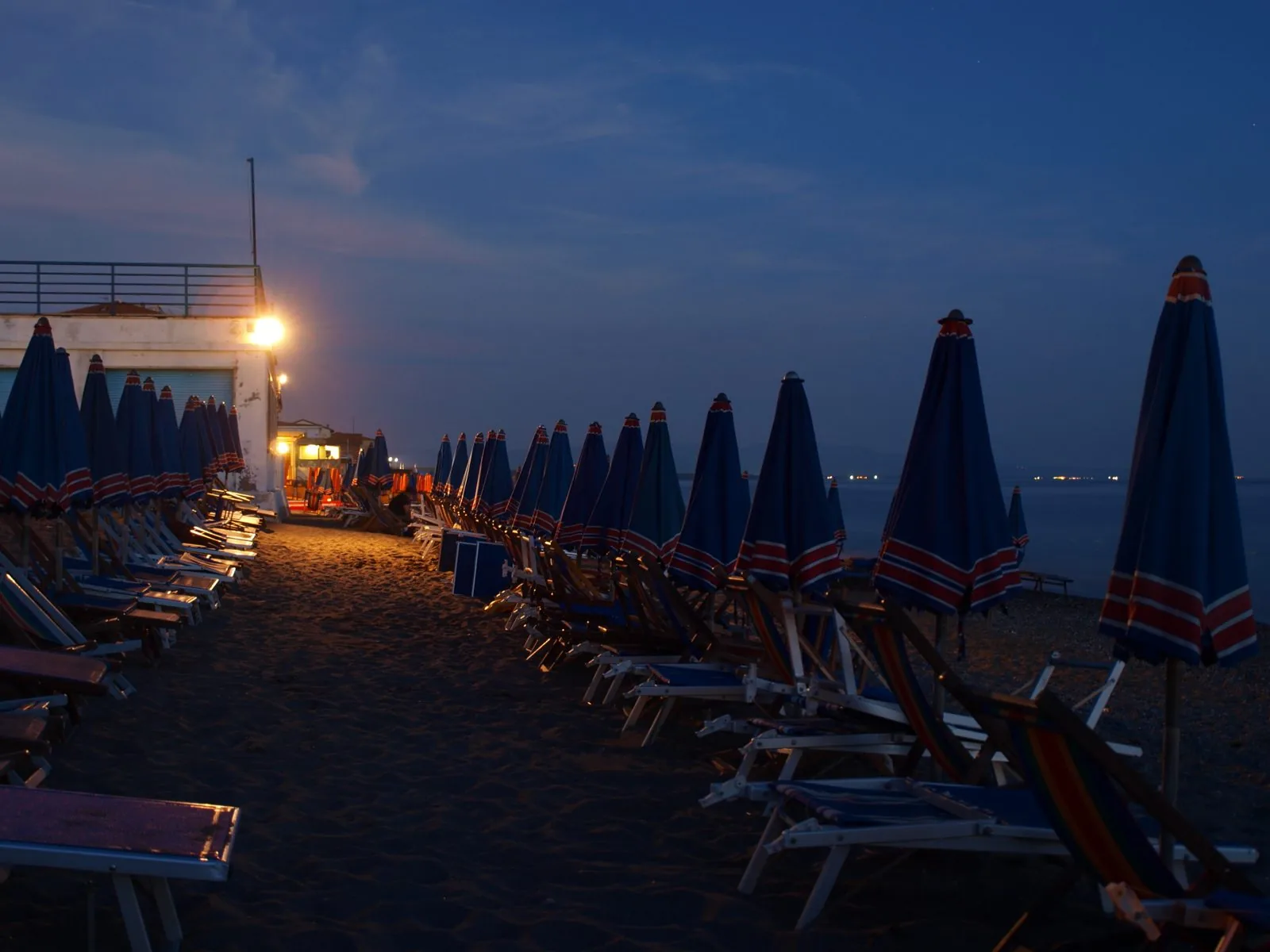 Ombrelloni chiusi e sdraio allineati sulla spiaggia di Marina di Cecina all'imbrunire, con le luci di uno stabilimento balneare